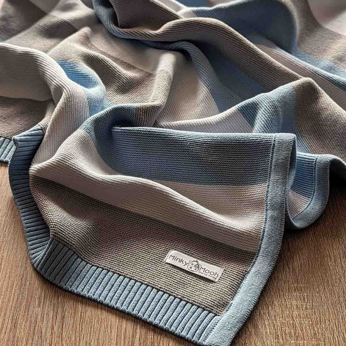 Detailansicht einer ausgebreiteten blau-grau-weißen Bio-Baumwolldecke auf einem Holzboden.