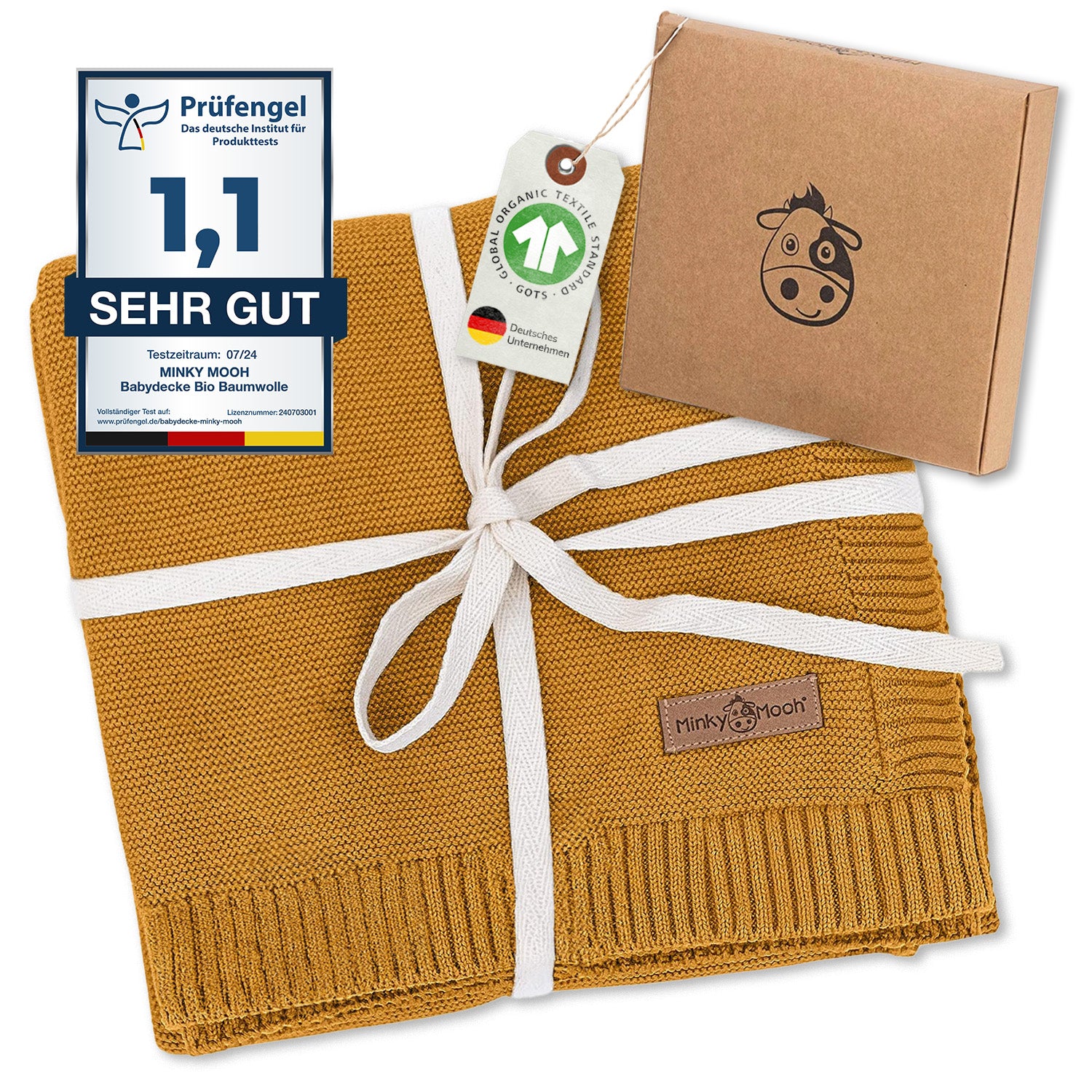 Senfgelbe Bio-Baumwolldecke für Babys mit Qualitätssiegel, Zertifizierungen und Verpackung. Gestrickte Decke mit weißer Schleife präsentiert. Nachhaltige Babyausstattung in Geschenkbox mit Kuh-Logo.