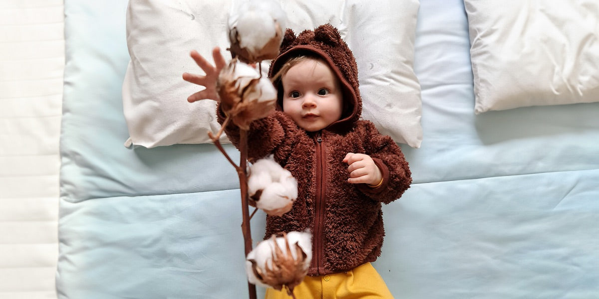 Ein Baby in einem braunen Bärenkostüm liegt auf einem Bett und greift nach Bio Baumwolle.