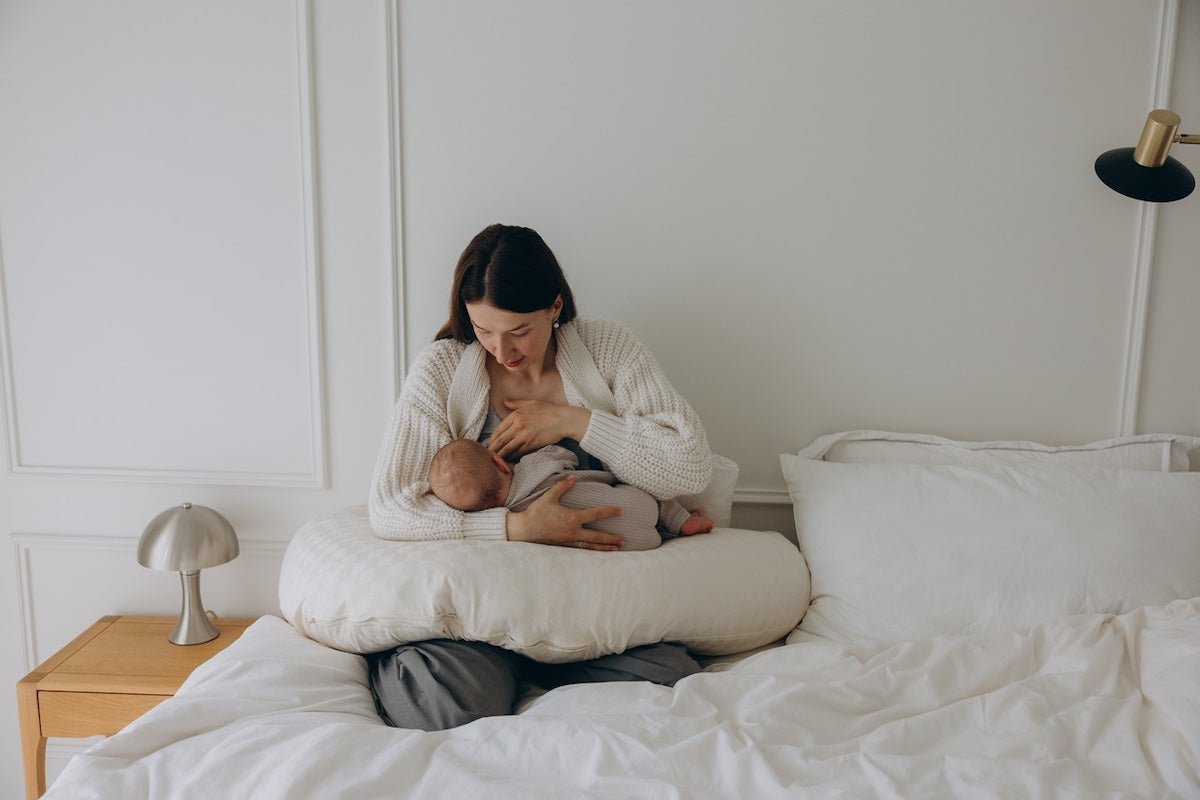 Eine Mutter stillt ihr Kind auf dem Bett, unterstützt von einem Bio-Stillkissen mit Kapokfüllung.