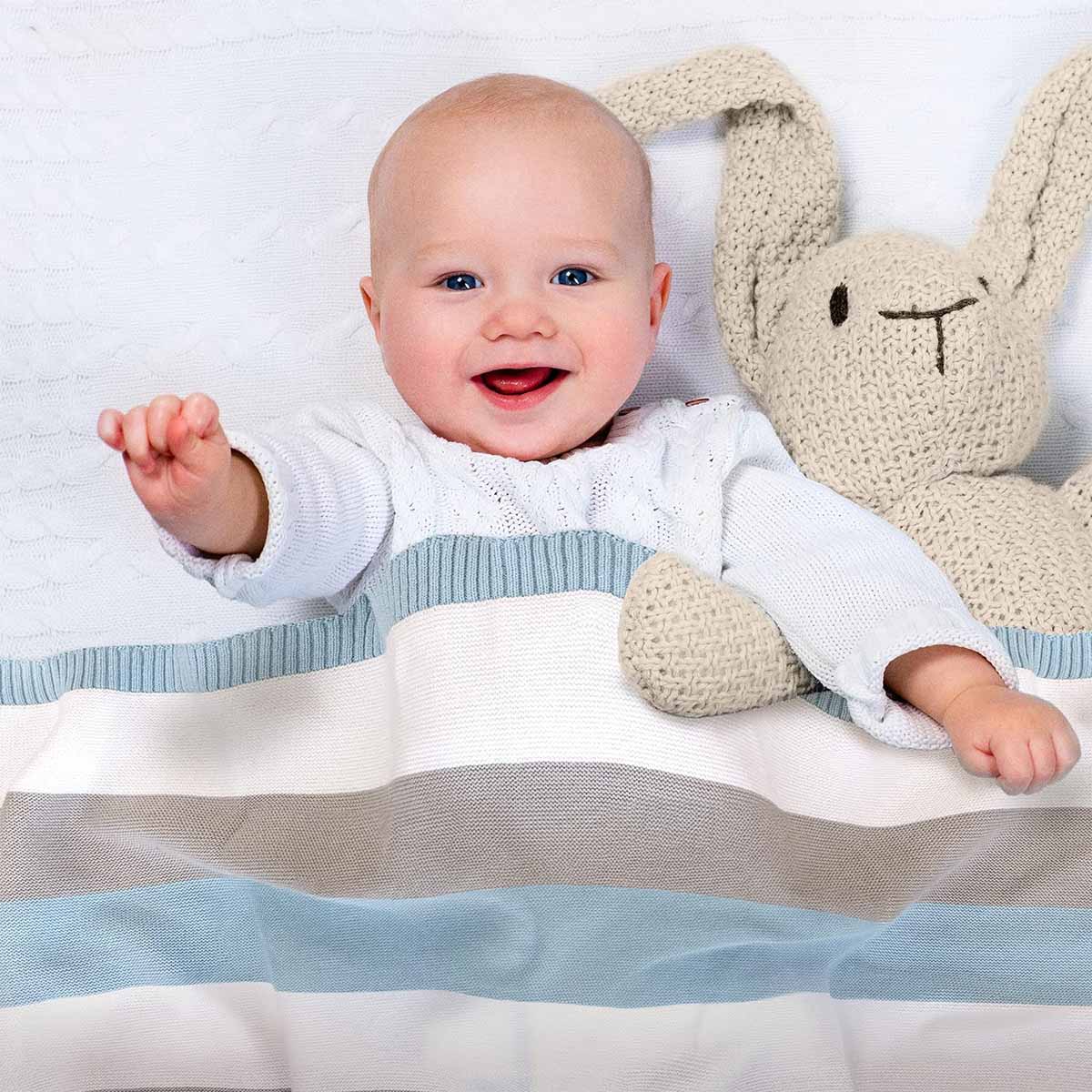 Glückliches Baby liegt im Bett, eingedeckt in blau-grau-weiß gestreifter Bio-Baumwolldecke. Es lächelt und winkt in die Kamera.