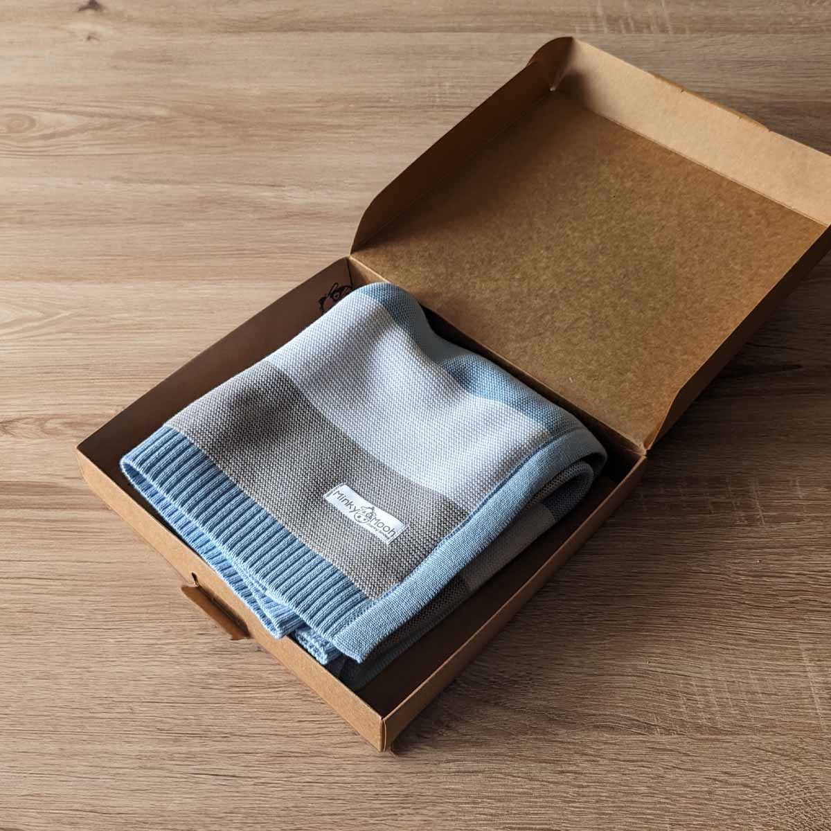 Gefaltete blaue und graue Bio-Baumwolldecke in einer offenen braunen Kartonverpackung.