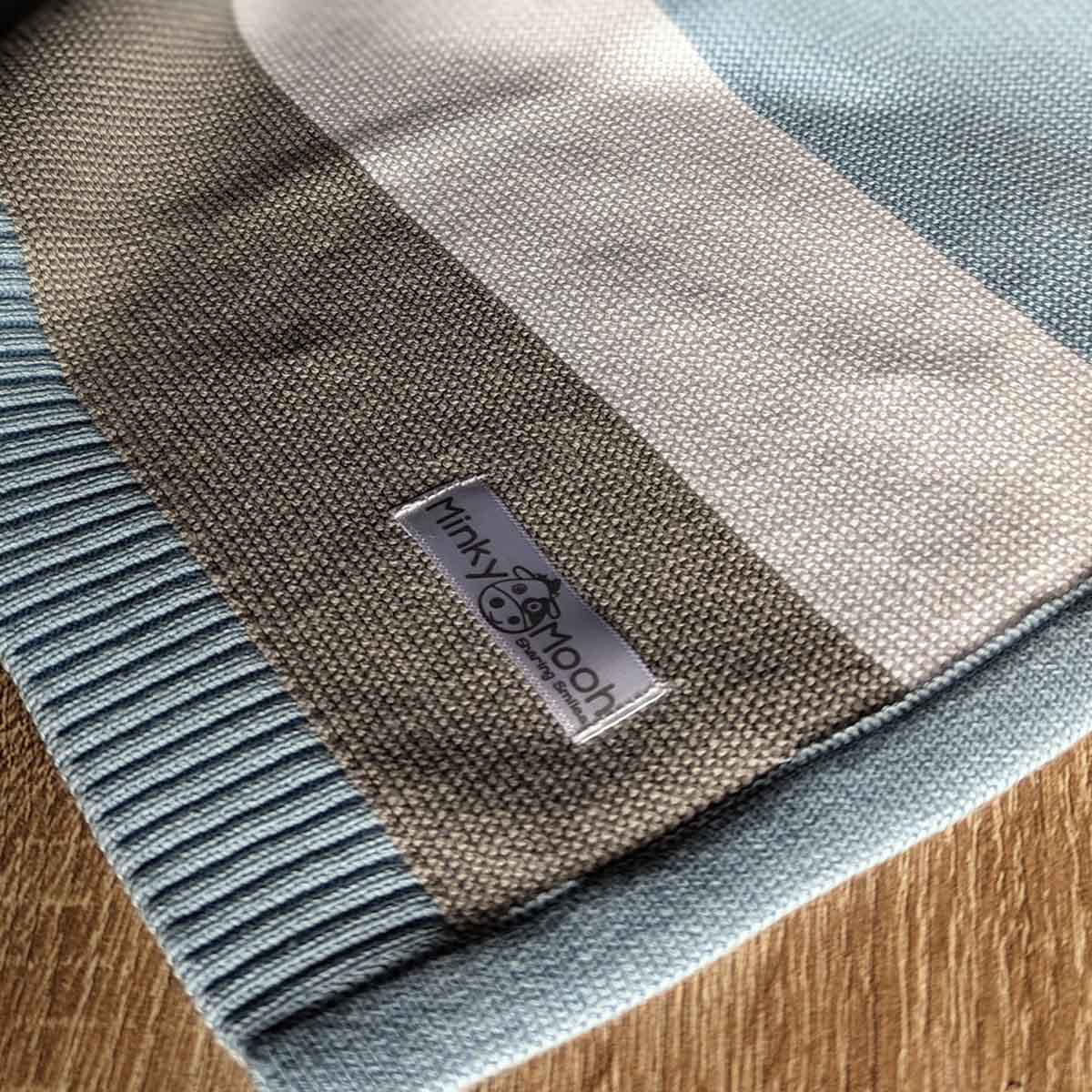 Nahaufnahme der Strick-Textur einer blau-weiß-grauen Bio-Baumwolldecke mit Logo-Etikett 'Minky Mooh'