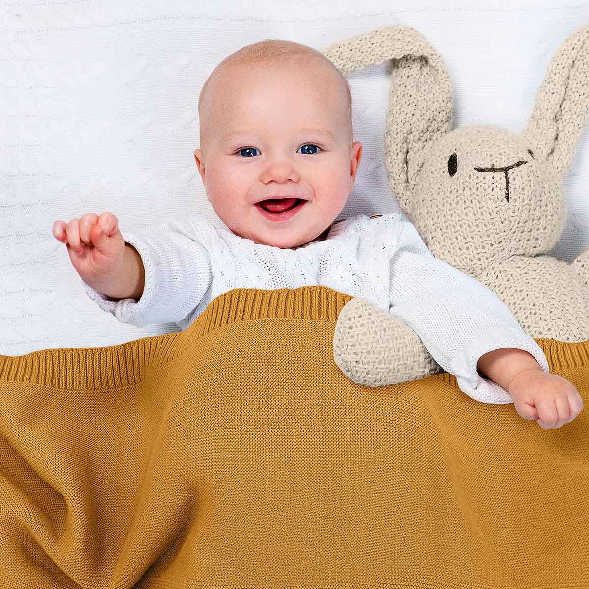 Glückliches Baby liegt im Bett, eingedeckt in dunkel-gelbe Bio-Baumwolldecke. Es lächelt und winkt in die Kamera.