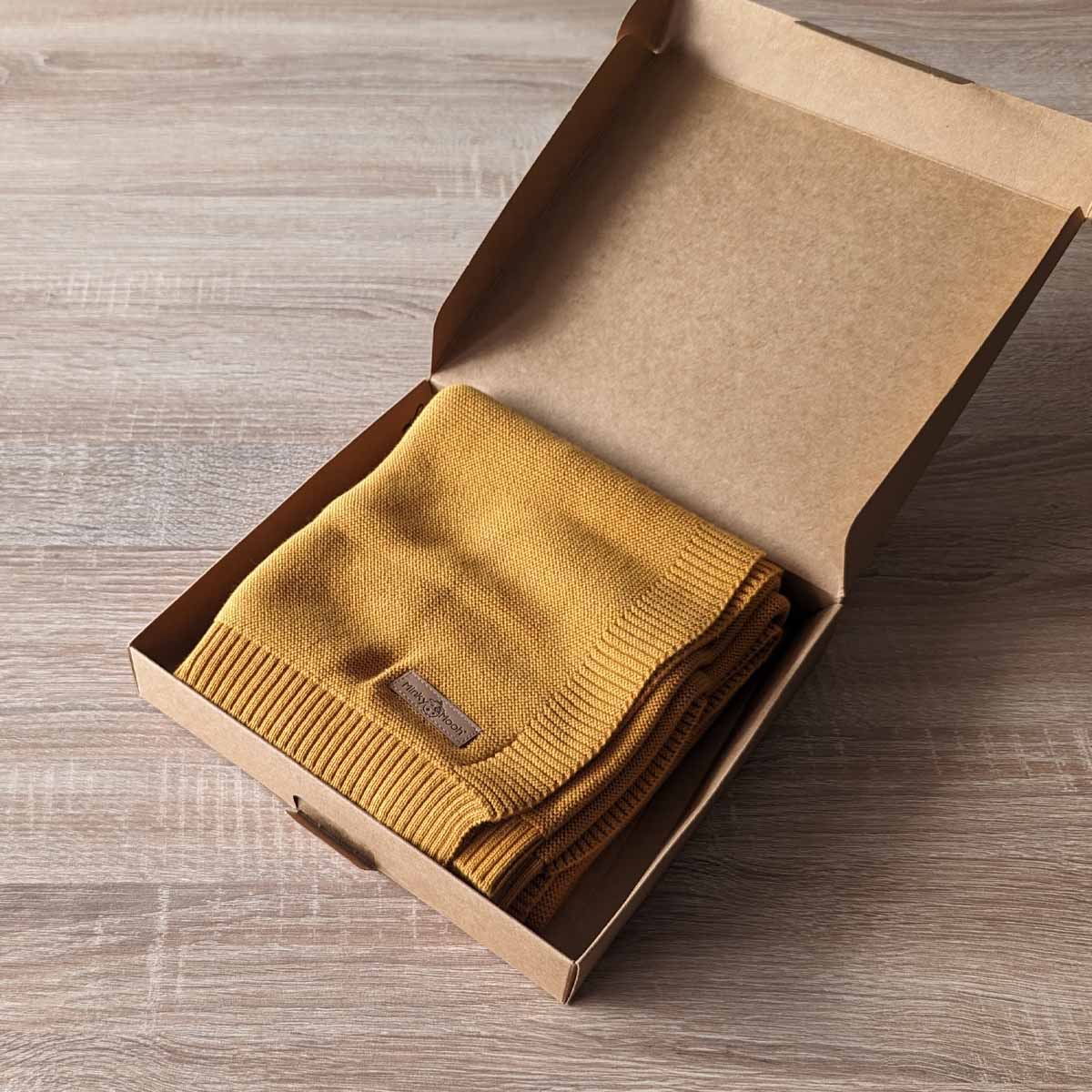 Gefaltete dunkel-gelbe Bio-Baumwolldecke in einer offenen braunen Kartonverpackung.