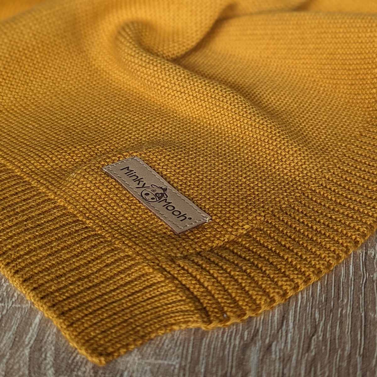 Nahaufnahme der Strick-Textur einer dunkel-gelbe Bio-Baumwolldecke mit Logo-Etikett 'Minky Mooh'.