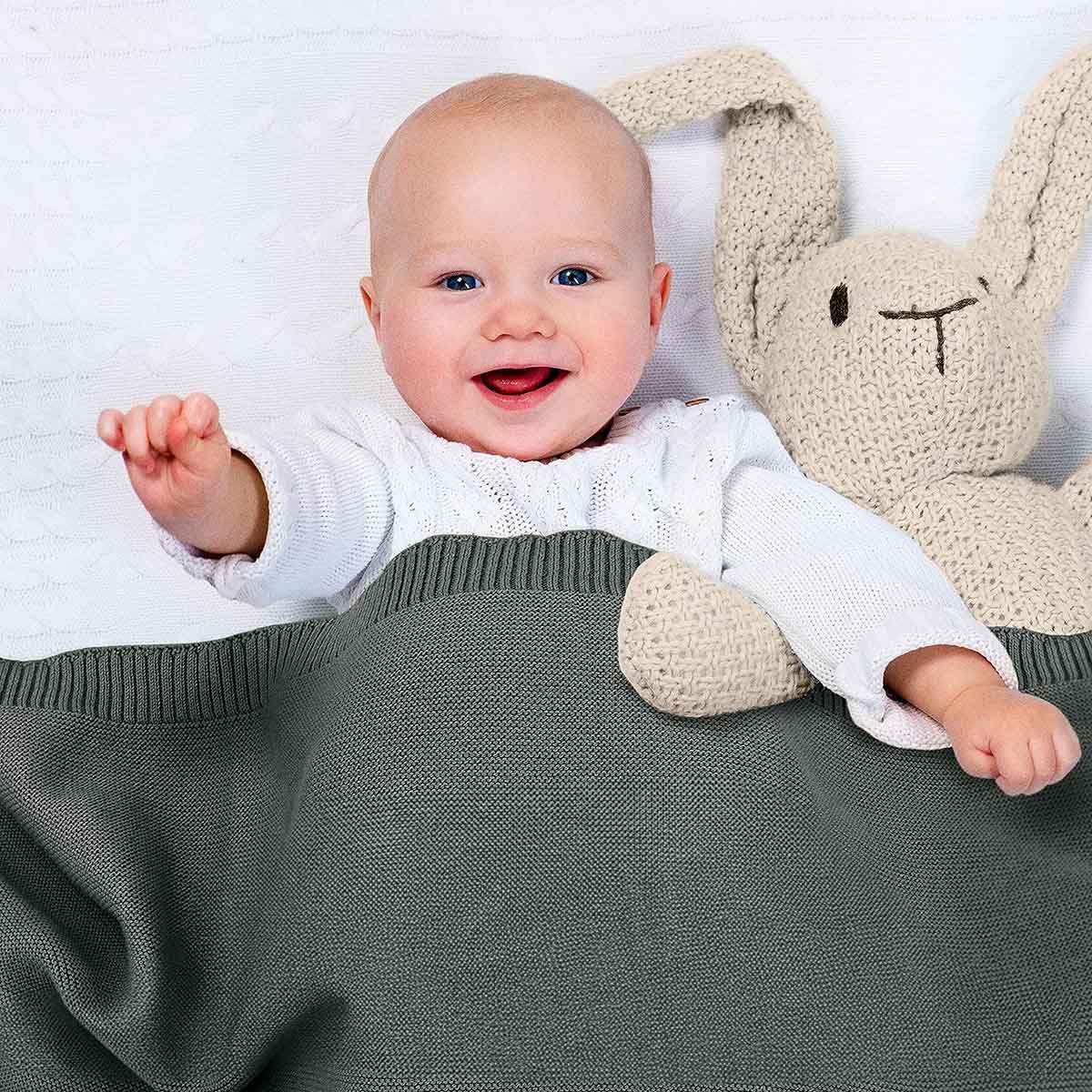 Glückliches Baby liegt im Bett, eingedeckt in dunkel-grüne Bio-Baumwolldecke. Es lächelt und winkt in die Kamera.