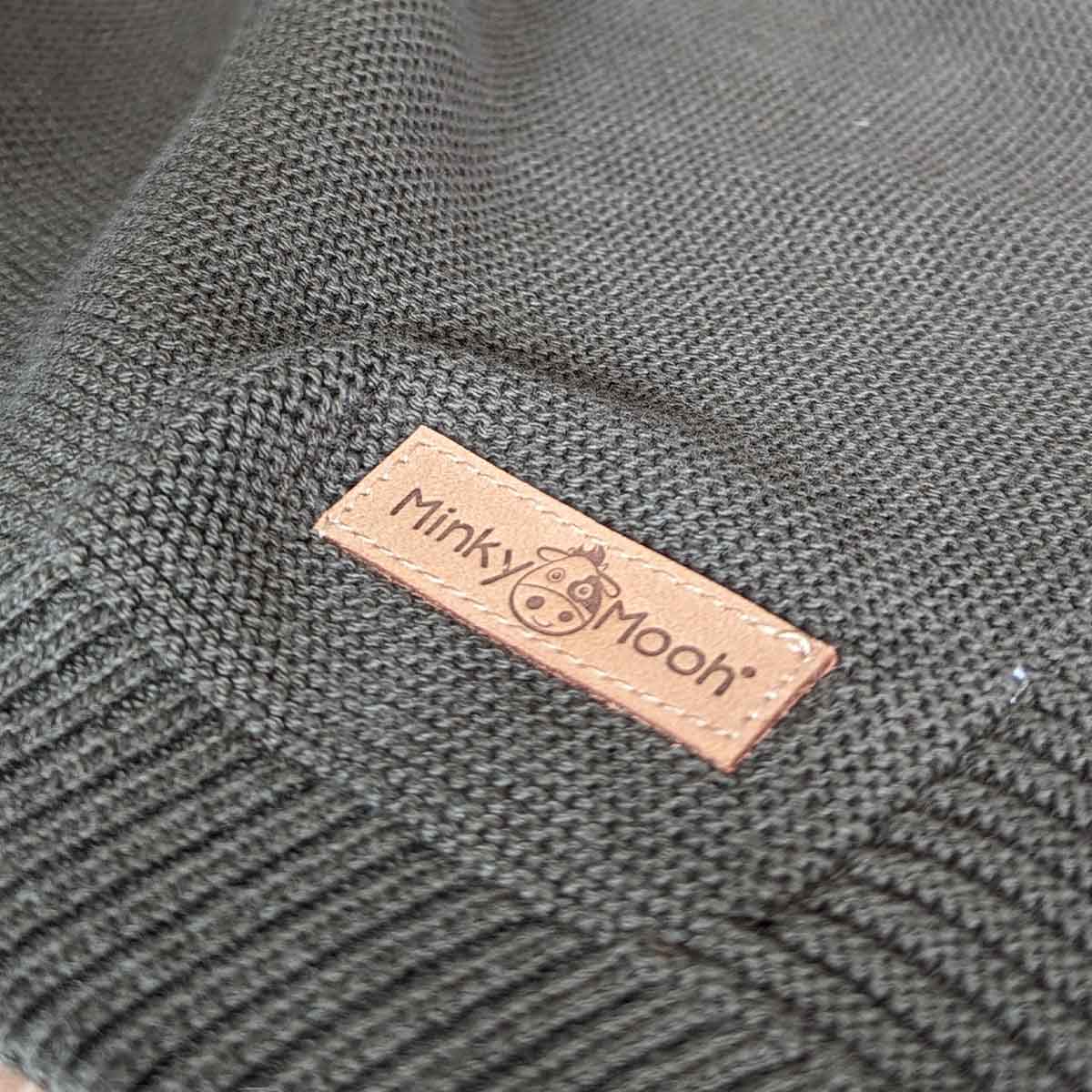 Nahaufnahme der Strick-Textur einer dunkel-grünen Bio-Baumwolldecke mit Logo-Etikett 'Minky Mooh'.