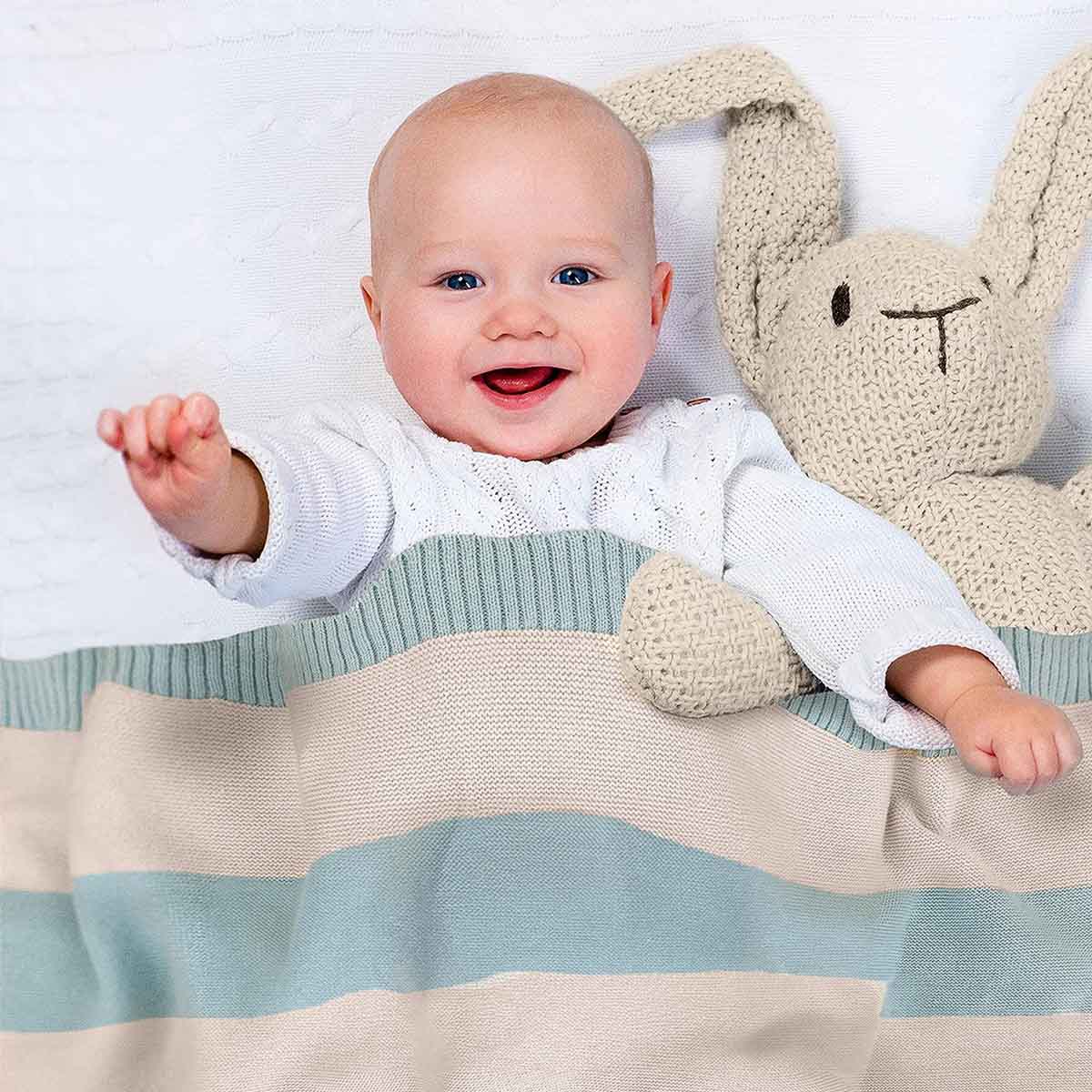 Glückliches Baby liegt im Bett, eingedeckt in mint-natur gestreifter Bio-Baumwolldecke. Es lächelt und winkt in die Kamera.
