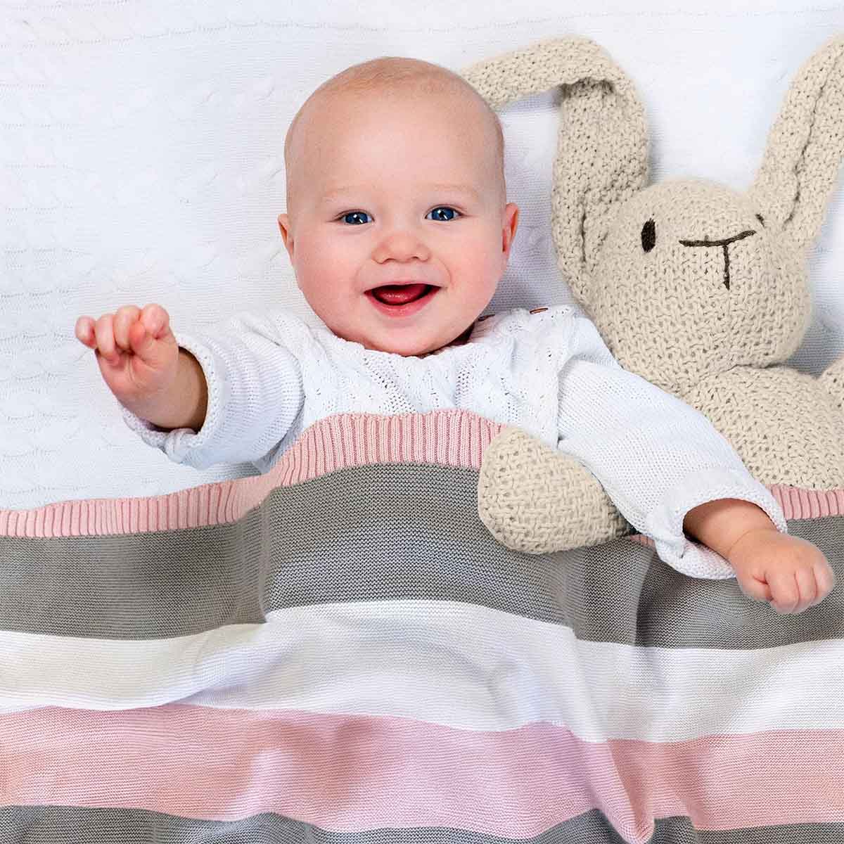 Glückliches Baby liegt im Bett, eingedeckt in rosa-grau-weiß gestreifter Bio-Baumwolldecke. Es lächelt und winkt in die Kamera.