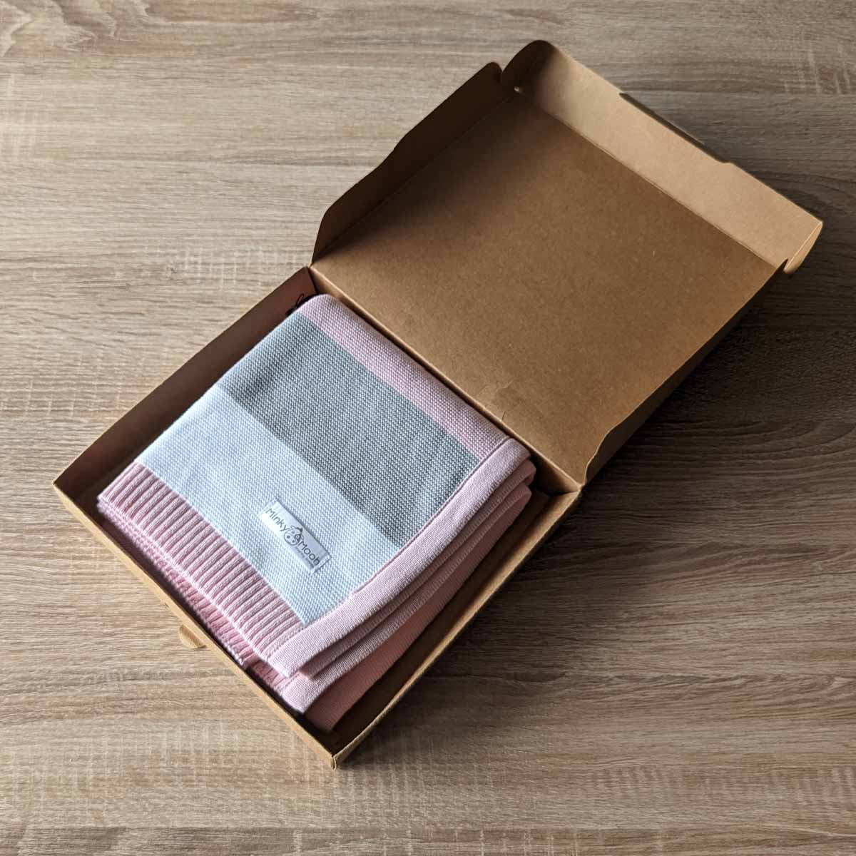 Gefaltete rosa-grau-weiß gestreifte Bio-Baumwolldecke in einer offenen braunen Kartonverpackung.