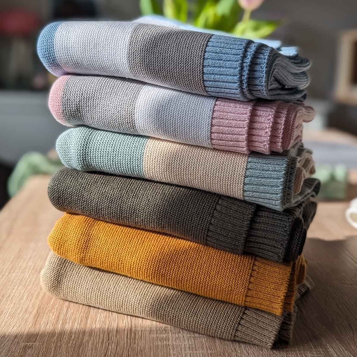 Stapel verschiedener gefalteter Bio-Baumwolldecken in mehreren Farben auf einem Holztisch.