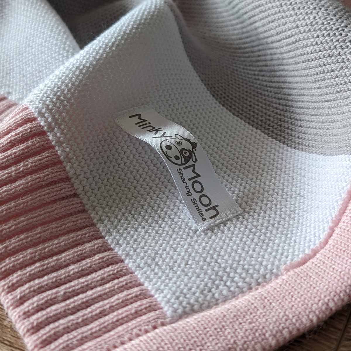 Nahaufnahme der Strick-Textur einer rosa-weiß-grauen Bio-Baumwolldecke mit Logo-Etikett 'Minky Mooh'