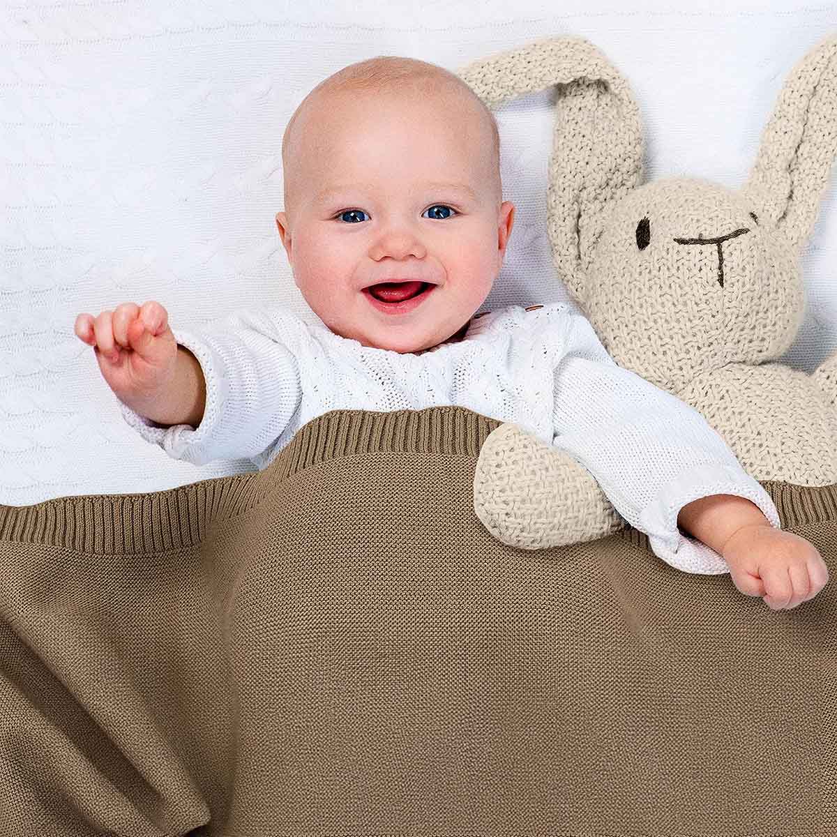 Glückliches Baby liegt im Bett, eingedeckt in taupe farbene Bio-Baumwolldecke. Es lächelt und winkt in die Kamera.