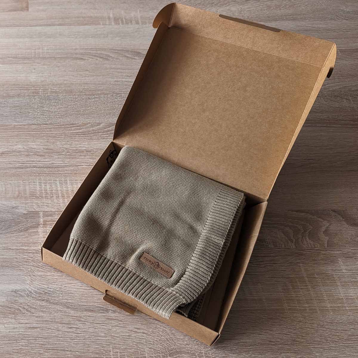 Gefaltete taupe farbene Bio-Baumwolldecke in einer offenen braunen Kartonverpackung.