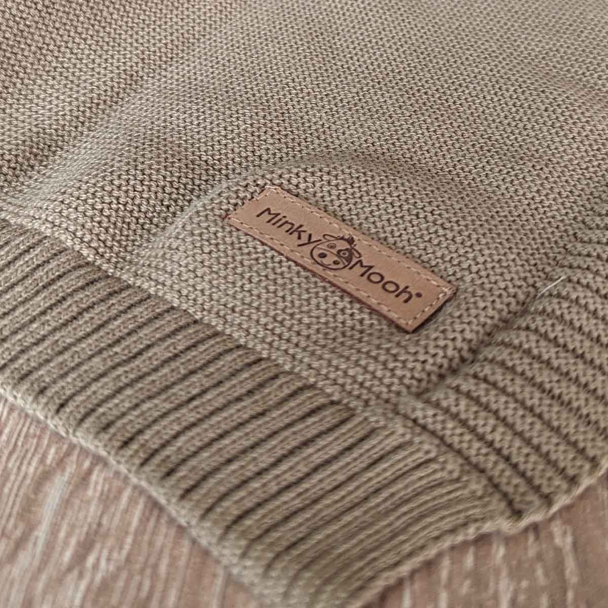 Nahaufnahme der Strick-Textur einer taupe farbenen Bio-Baumwolldecke mit Logo-Etikett 'Minky Mooh'